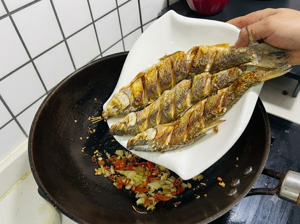 炒めたニンニク、生姜、唐辛子が入った中華鍋へ、白い皿から3尾の揚げた魚を戻している様子。