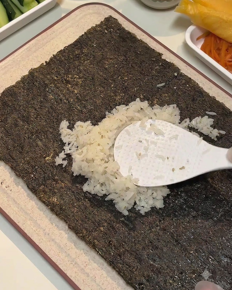 白いご飯のしゃもじを使って、味付けしたご飯を海苔の中央部分に均一に広げている様子。