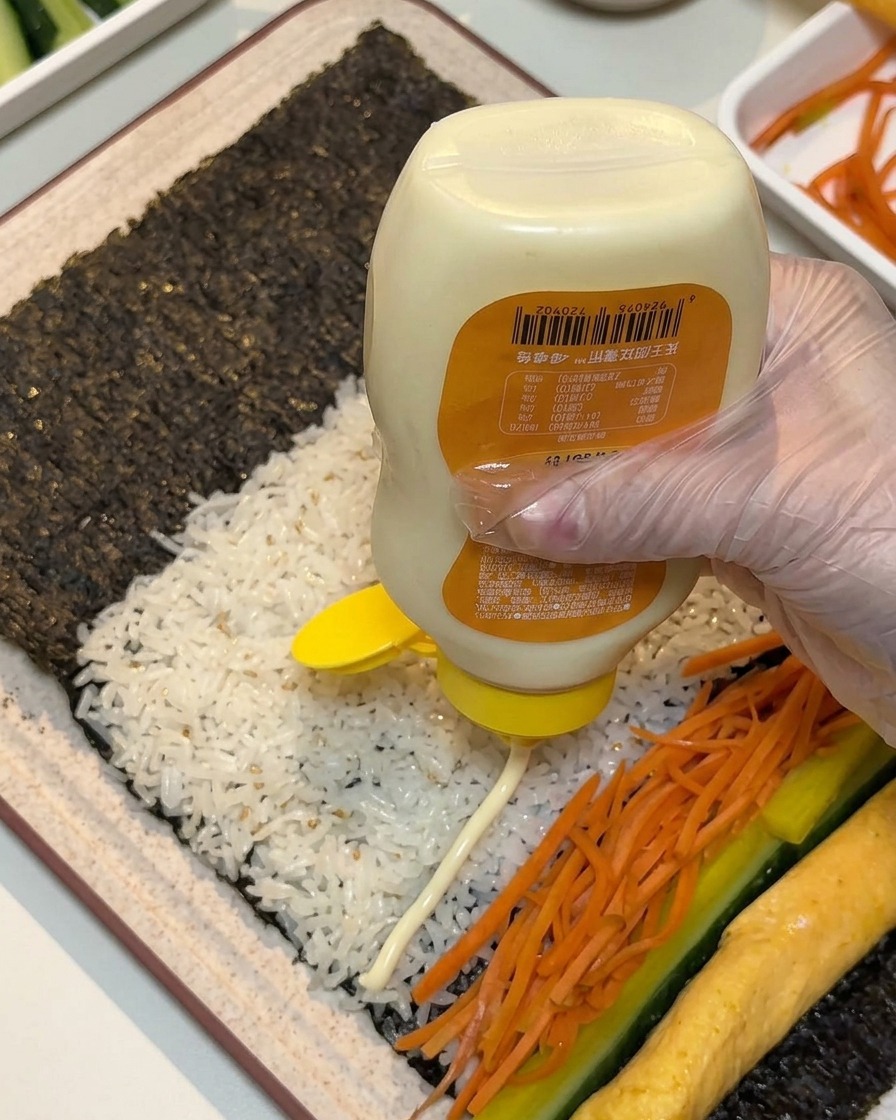 手袋を着用した手が、海苔とご飯の上に並べられた具材の上に向けて、ボトルから白いマヨネーズを水平に絞り出している様子。