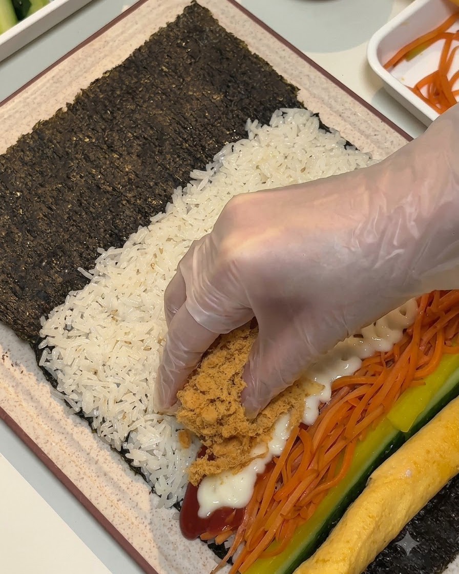 手袋を着用した手が、海苔の上の具材の上に茶色のふわふわした肉でんぶとランチョンミートの細切りを乗せている様子。