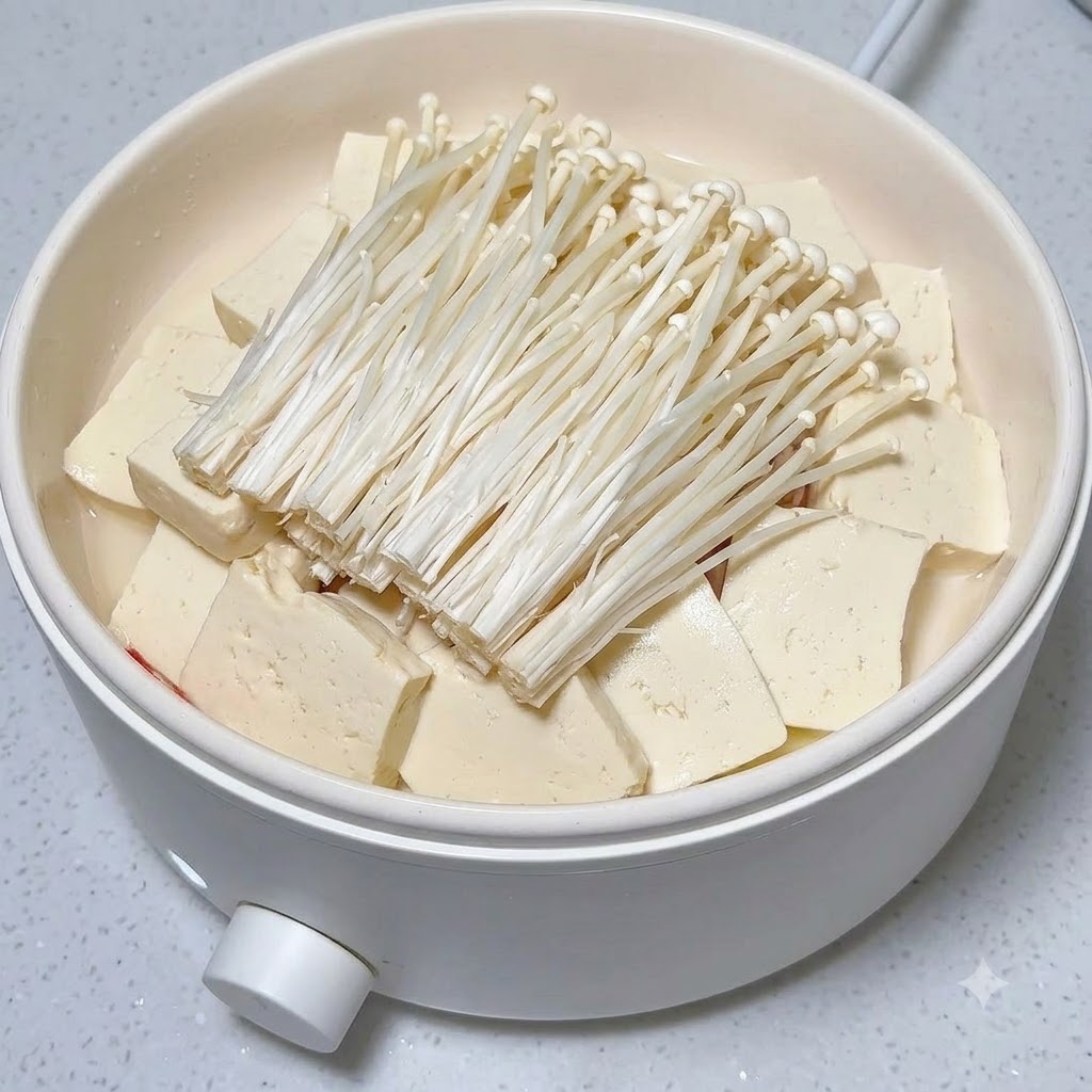 白い鍋の中のエノキと豆腐の山の上に、新鮮な赤唐辛子の輪切りがきれいに並べられている様子。