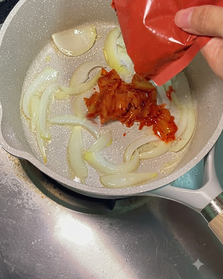 炒めた玉ねぎが入った熱いフライパンに、赤いパッケージから鮮やかな赤い刻みキムチを加えている様子。