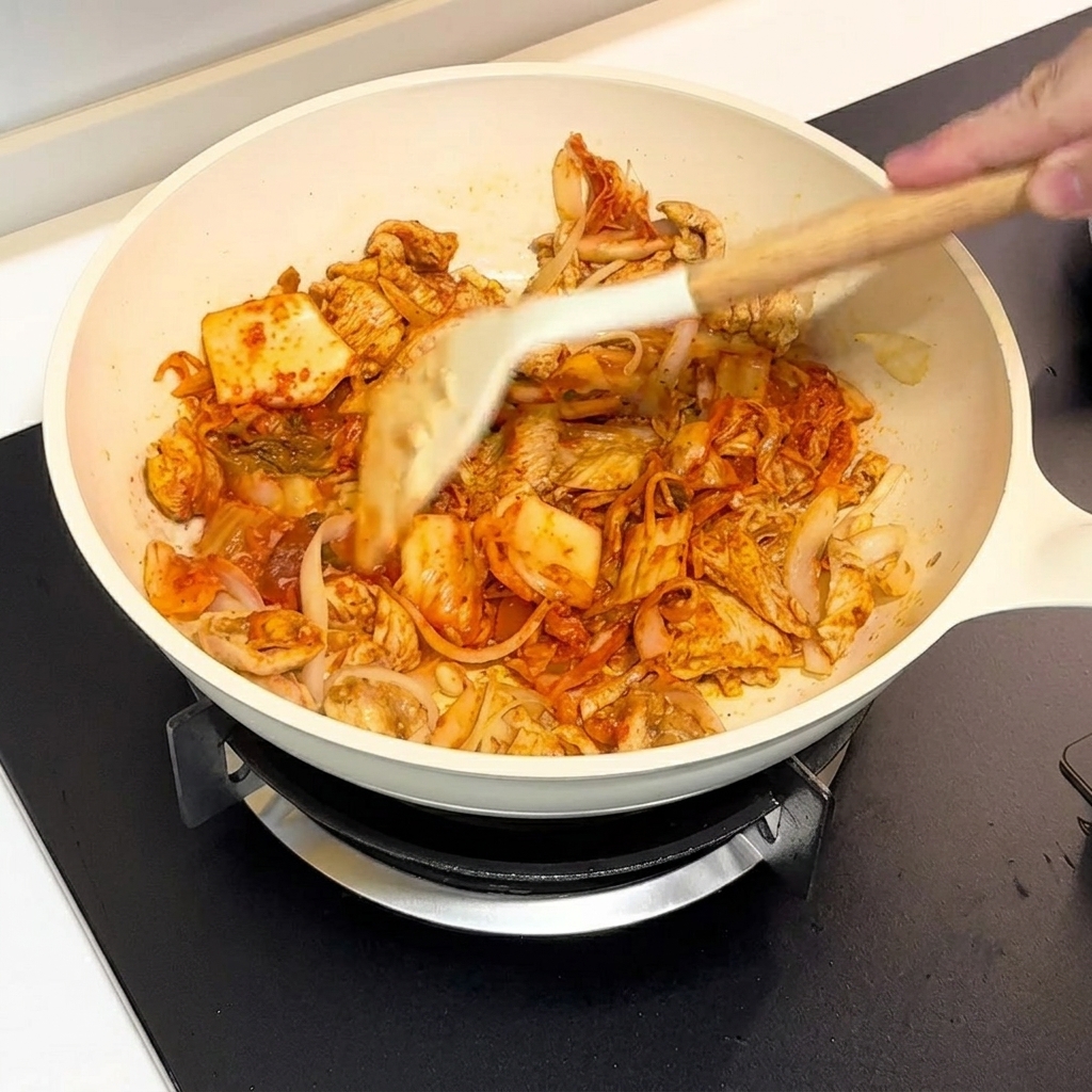 白いフライパンの中で、スパチュラを使ってキムチと鶏肉を一緒に炒めている様子。