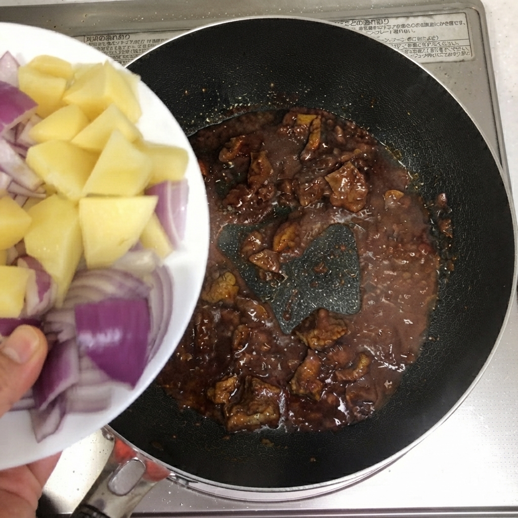 お皿からスライスしたジャガイモと赤玉ねぎが、牛肉の入ったフライパンに投入されている様子