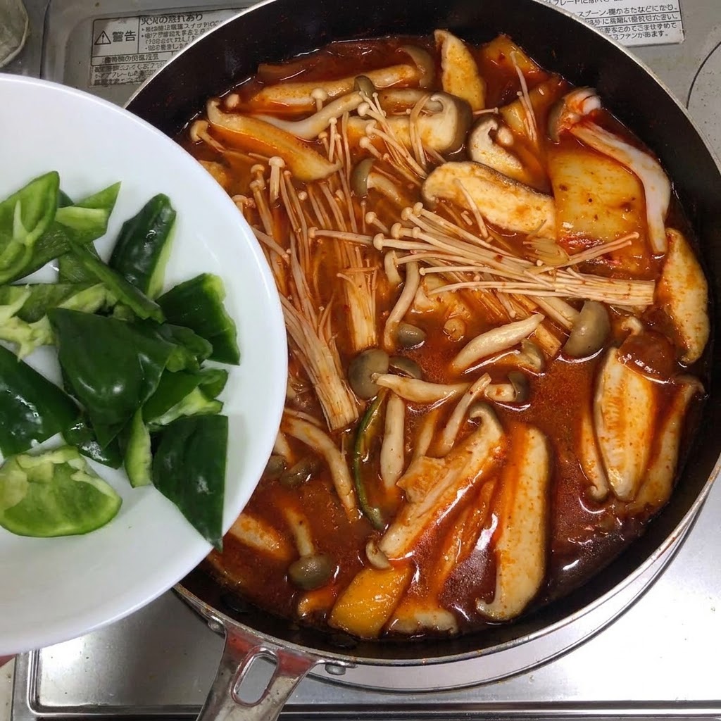 白いお皿からスライスした青唐辛子が、真っ赤なキムチスープの鍋に加えられている様子