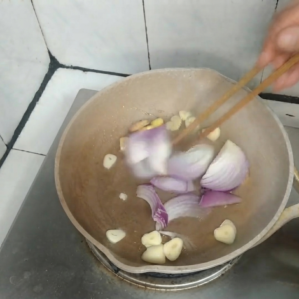 フライパンの中で、木製のお箸を使ってスライスした赤玉ねぎ、にんにく、生姜を一緒に炒めている様子。