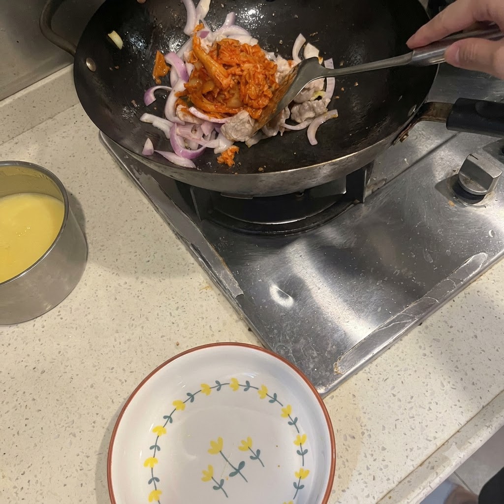 炒めた豚バラ肉と赤玉ねぎが入った中華鍋に、ヘラを使って真っ赤なキムチを入れている様子。