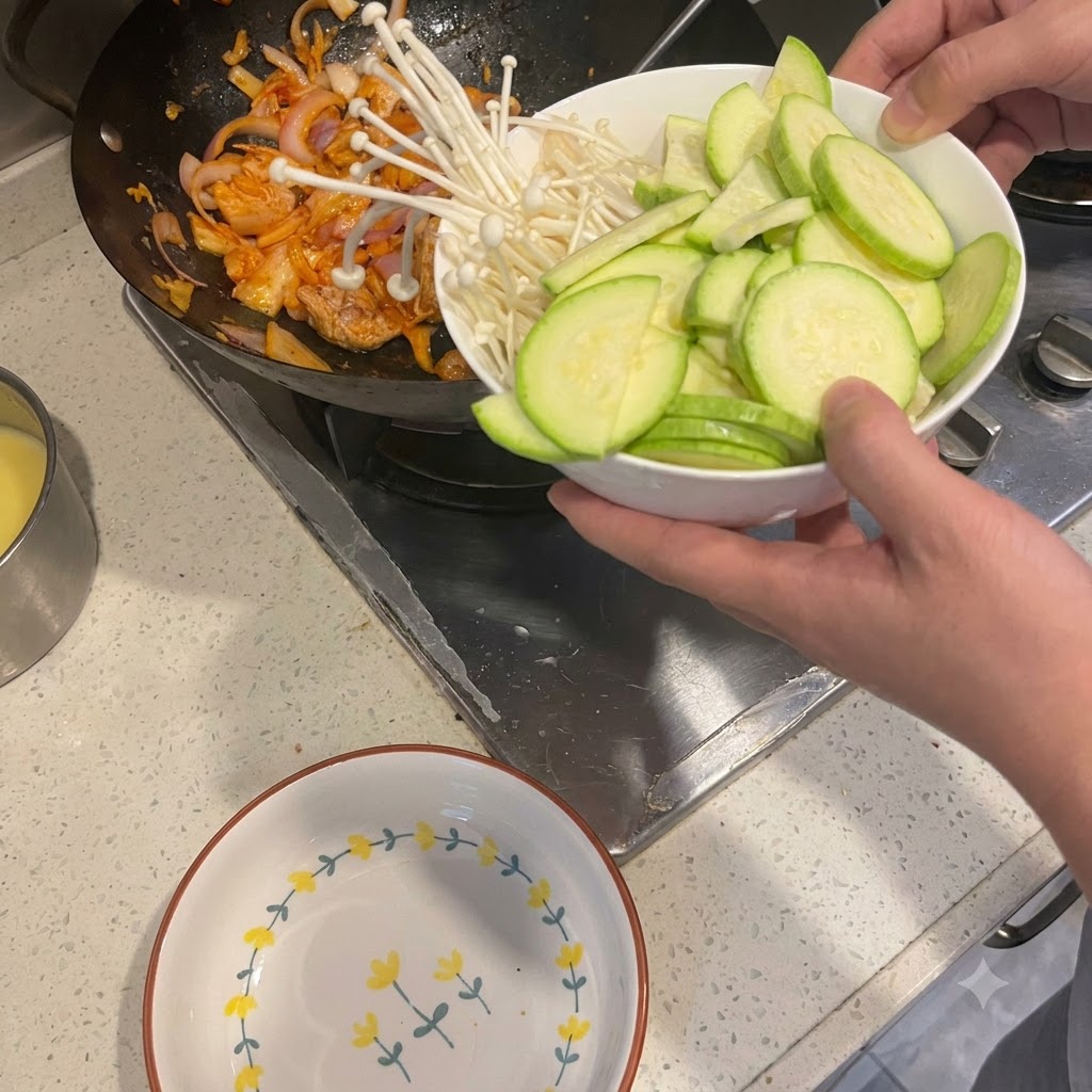 白いボウルから、炒めた豚肉と玉ねぎが入った中華鍋にスライスしたズッキーニとエノキタケを移しているところ。