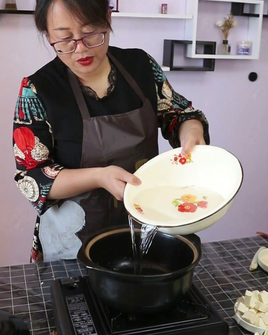 ポータブルコンロに置かれた黒い土鍋に、装飾的なボウルからきれいな水を注いでいる様子。