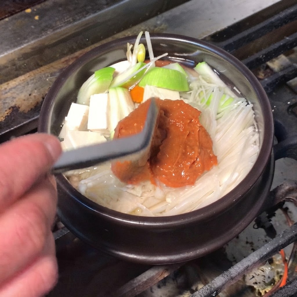 シリコンヘラを使い、コクのある茶色い味噌と赤いコチュジャンを調理鍋に加えている様子。