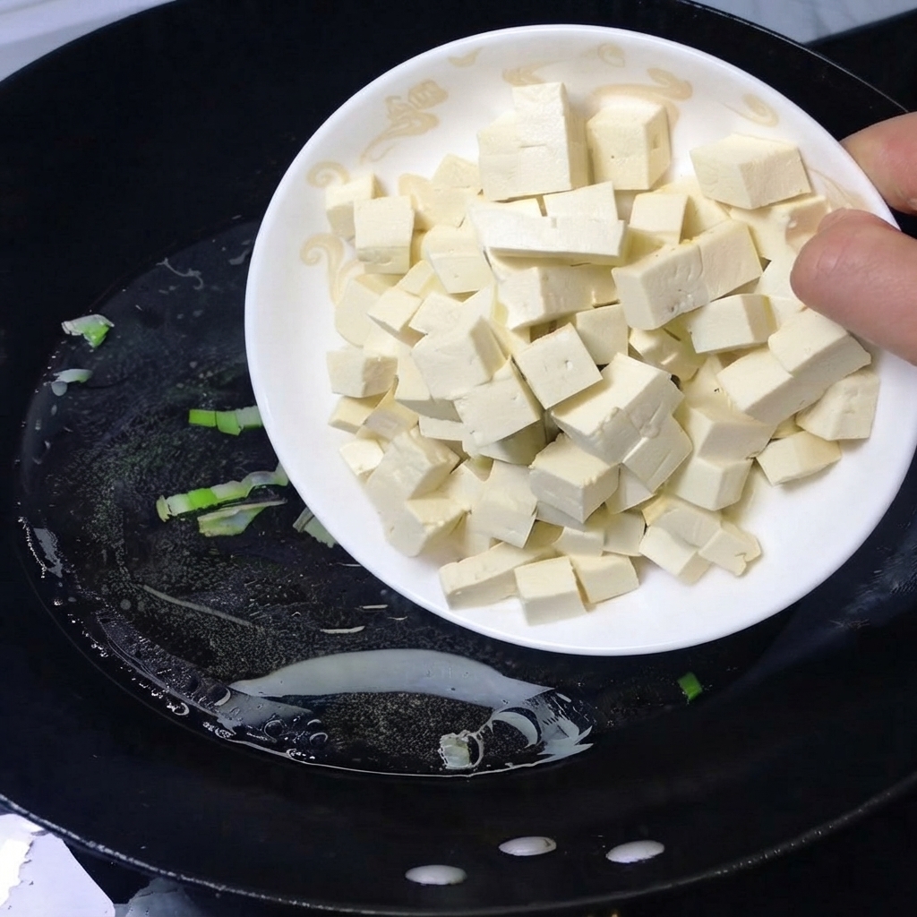角切りの白い豆腐を乗せた皿から、沸騰したお湯が入った黒い中華鍋に豆腐を滑り込ませている様子