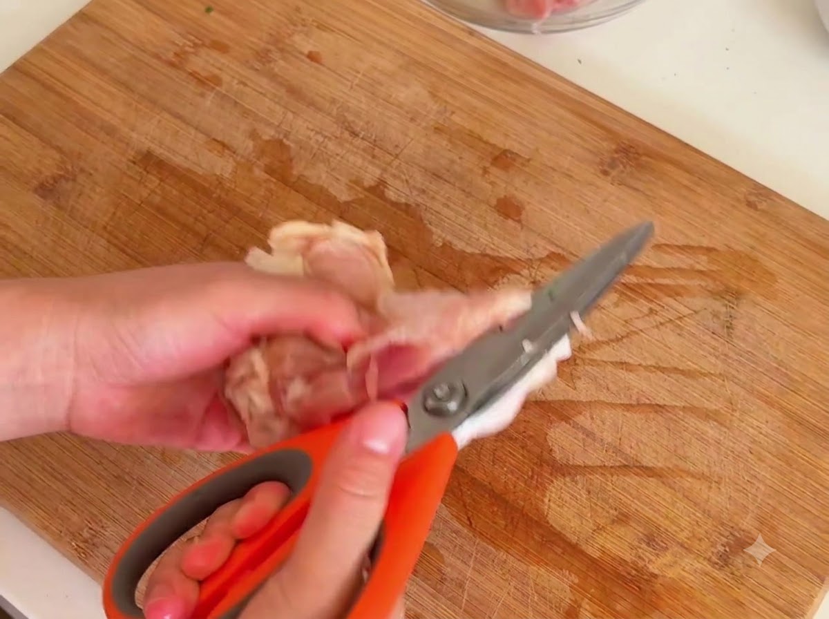 木製のまな板の上で、オレンジ色のキッチンバサミを使い、生の鶏もも肉の骨から肉を切り離しているところ。