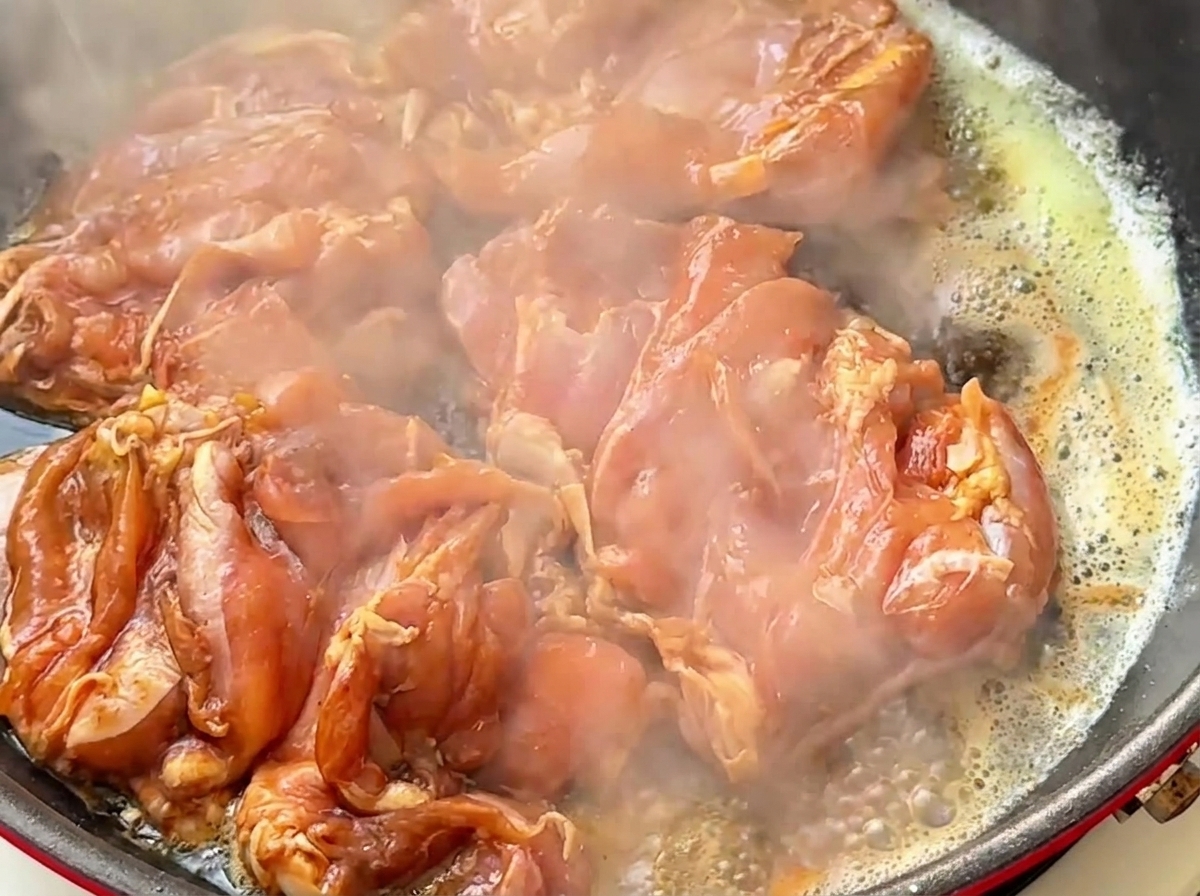 バターを引いた熱々のフライパンの中で、マリネされた生肉の鶏もも肉がジューシーな音を立てて湯気を上げているところ。