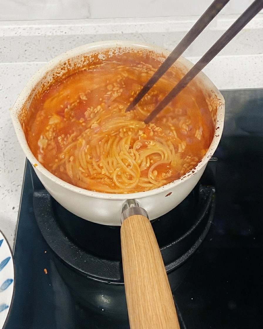 コンロの上で煮立っているトマトベースのパスタの鍋。ソースにとろみをつけるために箸で麺をかき混ぜている。