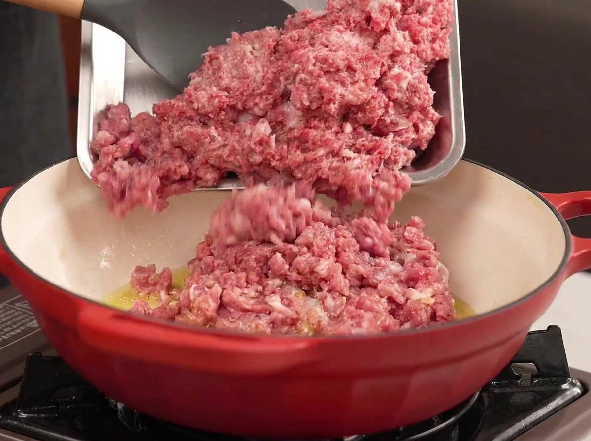 金属製のトレイから、熱した油を入れた赤い鍋に牛ひき肉と豚ひき肉を入れている様子。