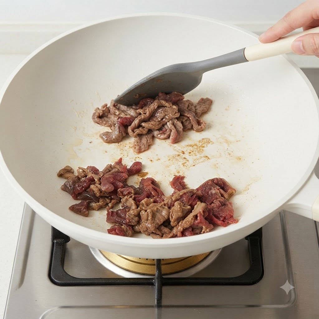 下味をつけた牛肉のスライスを白いフライパンで灰色のシリコンヘラを使って炒めている様子