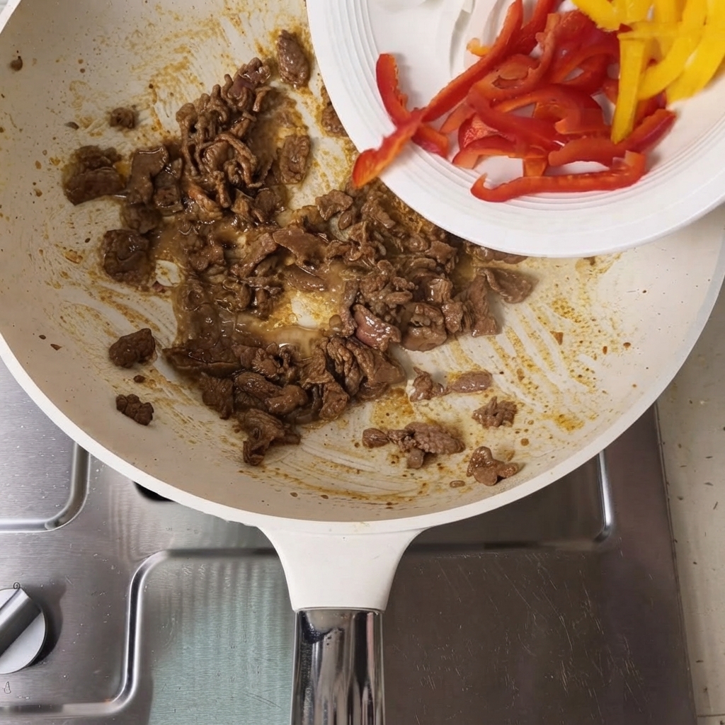 スライスした赤と黄色のパプリカが白い皿から調理済みの牛肉が入ったフライパンに注がれている様子