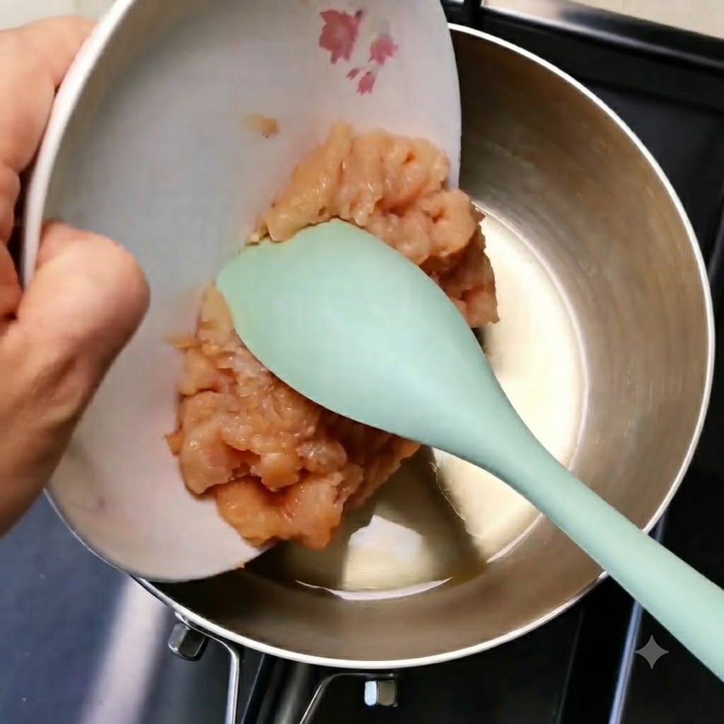 緑色のスパチュラを使い、白いお皿から金属製のフライパンへと鶏ひき肉を滑り込ませている様子。