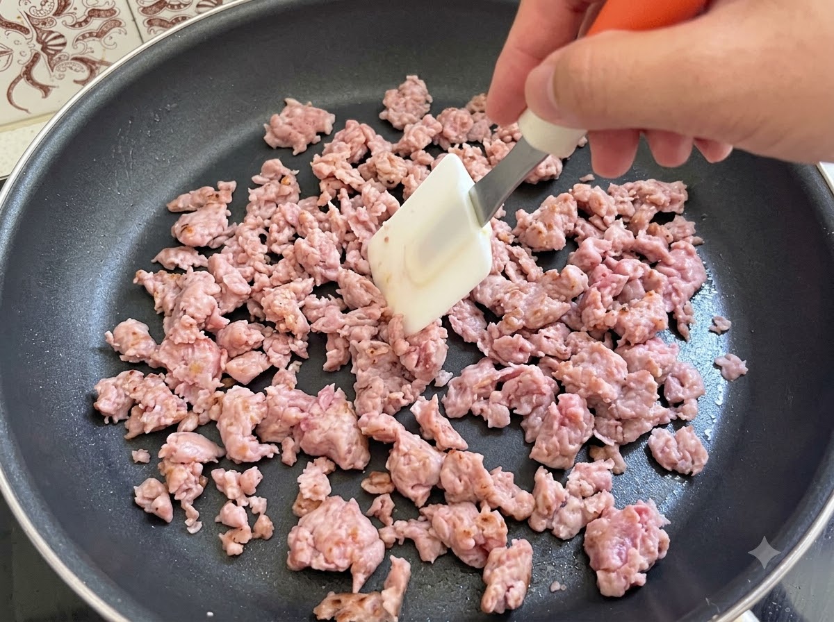 フライパンの中で、スパチュラを使ってイタリアンソーセージ肉が炒められ、かき混ぜられている様子。