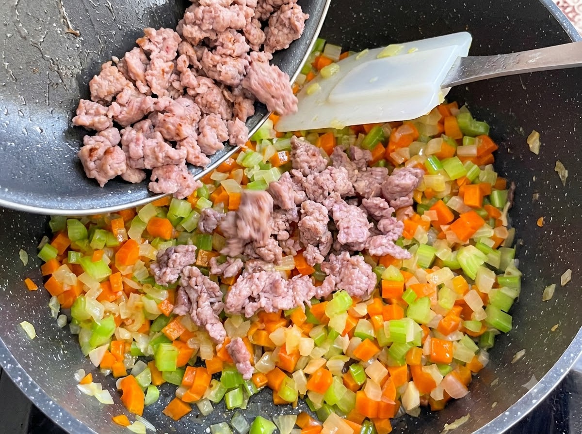 炒めたソーセージ肉がフライパンから野菜の入った鍋に注ぎ入れられ、混ぜ合わされている様子。