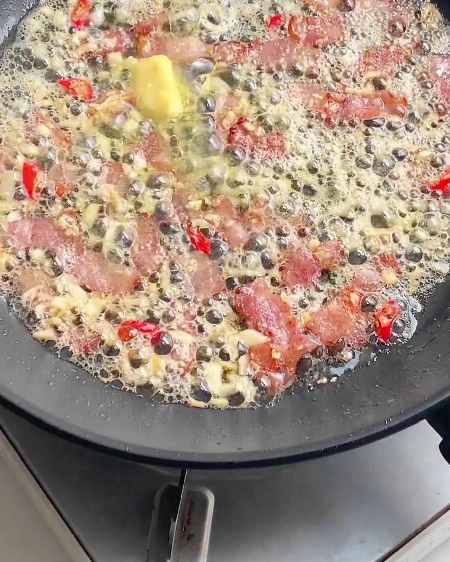 茹で上がったパスタを、ベーコン、溶かしバター、みじん切りニンニク、赤唐辛子の入った熱々のフライパンに投入している様子。