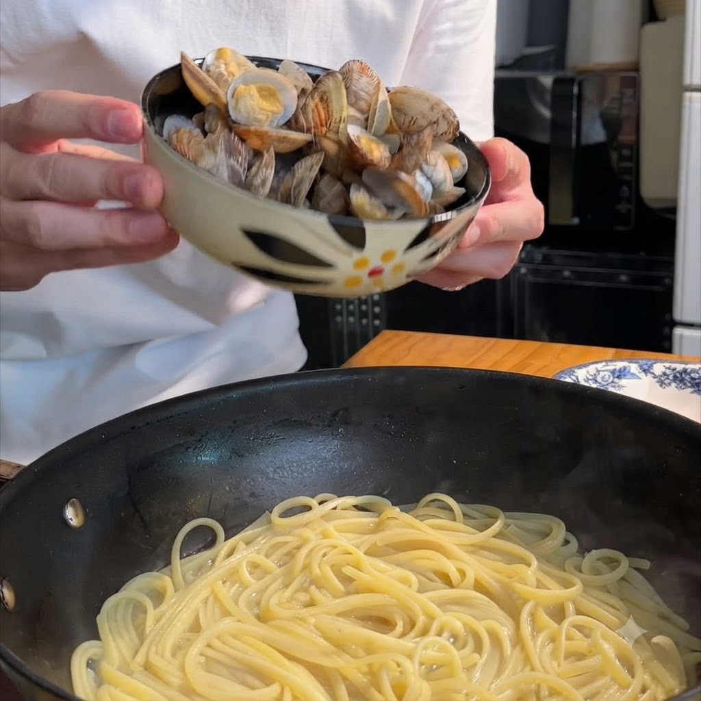 柄付きのボウルからスパゲッティの入ったフライパンに調理済みのハマグリが移されているところ。
