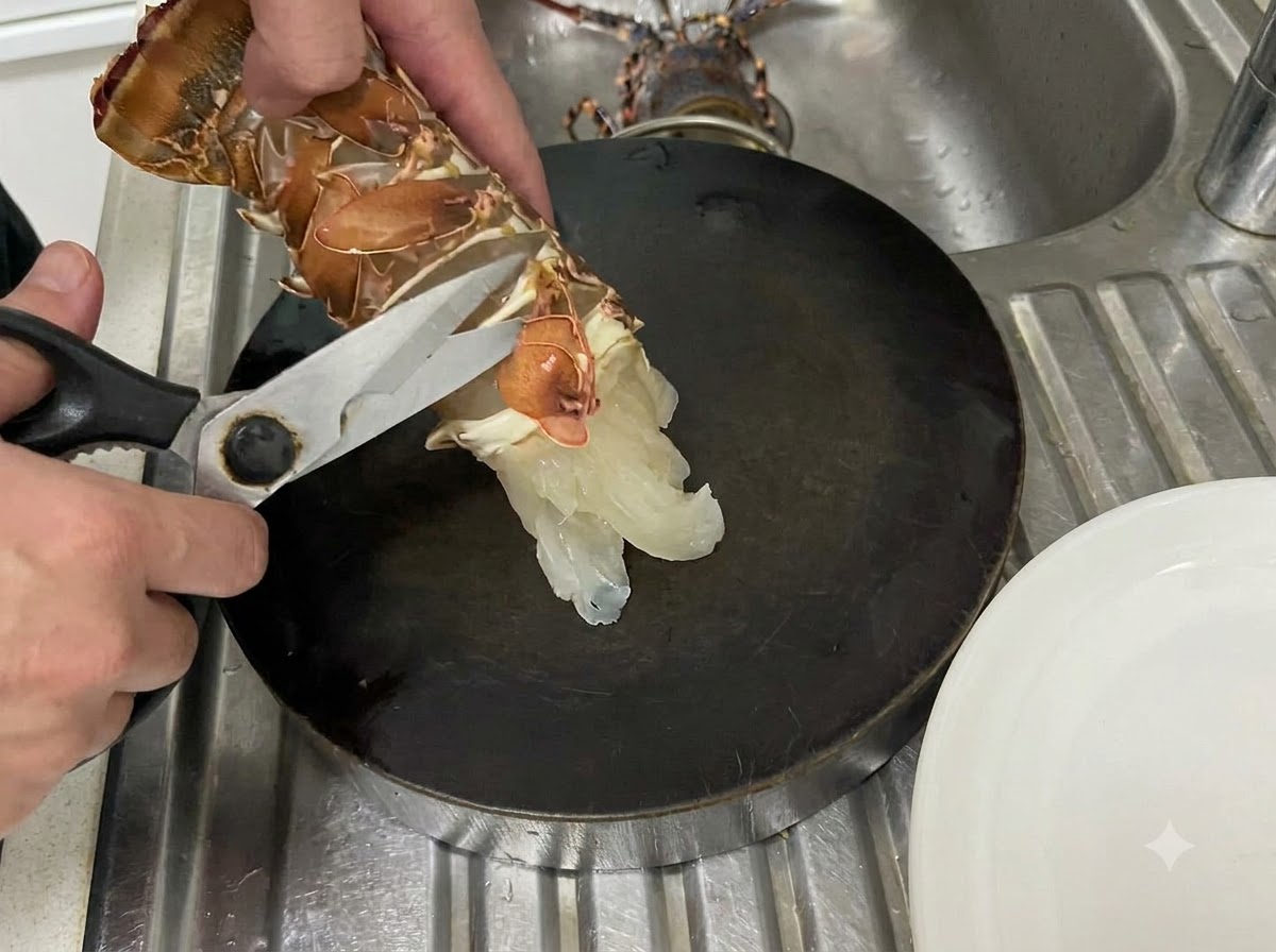 暗いまな板の上で、キッチンスパチュラを使い生きたロブスターの尾の裏側の薄皮に沿ってハサミを入れている手。