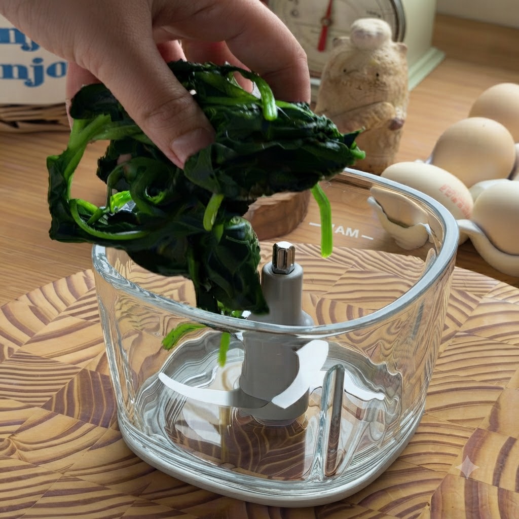 水気を絞った茹でほうれん草の束を、空のガラス製フードプロセッサーのボウルに入れている手。