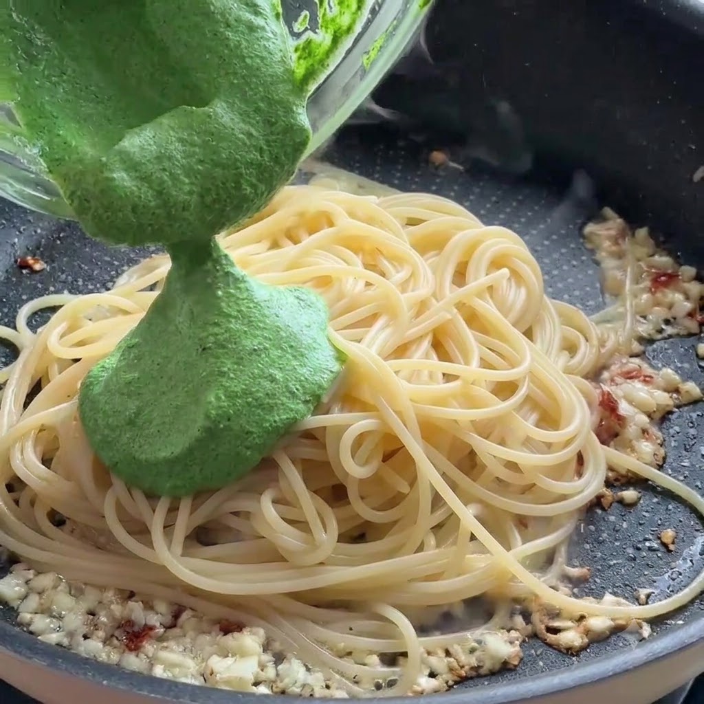 フライパンの中のスパゲッティの上に、ガラスボウルから緑色のペストソースを注いでいるところ。