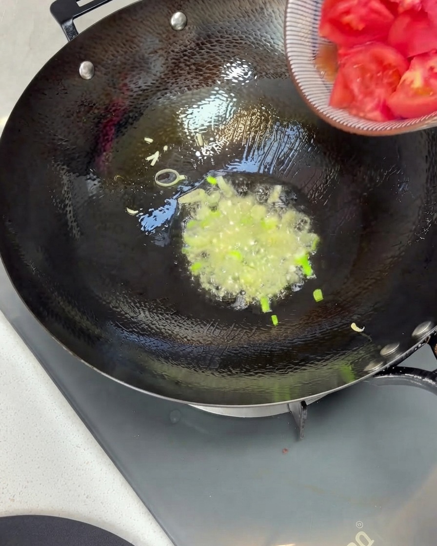 ボウルからざく切りにしたトマトを、青ねぎと油の入った中華鍋に投入している様子