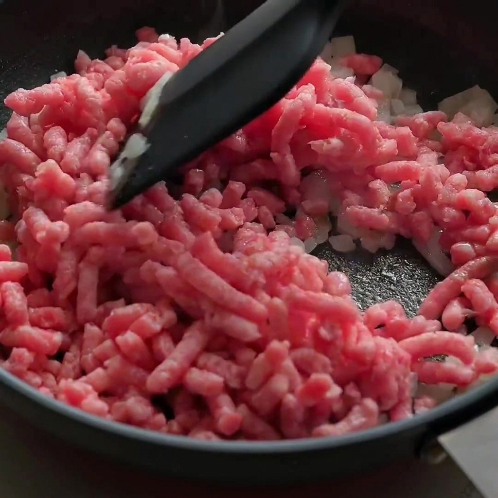 黒いフライパンの中で、生のピンク色の牛ひき肉をほぐしながら玉ねぎと一緒に炒めている様子。