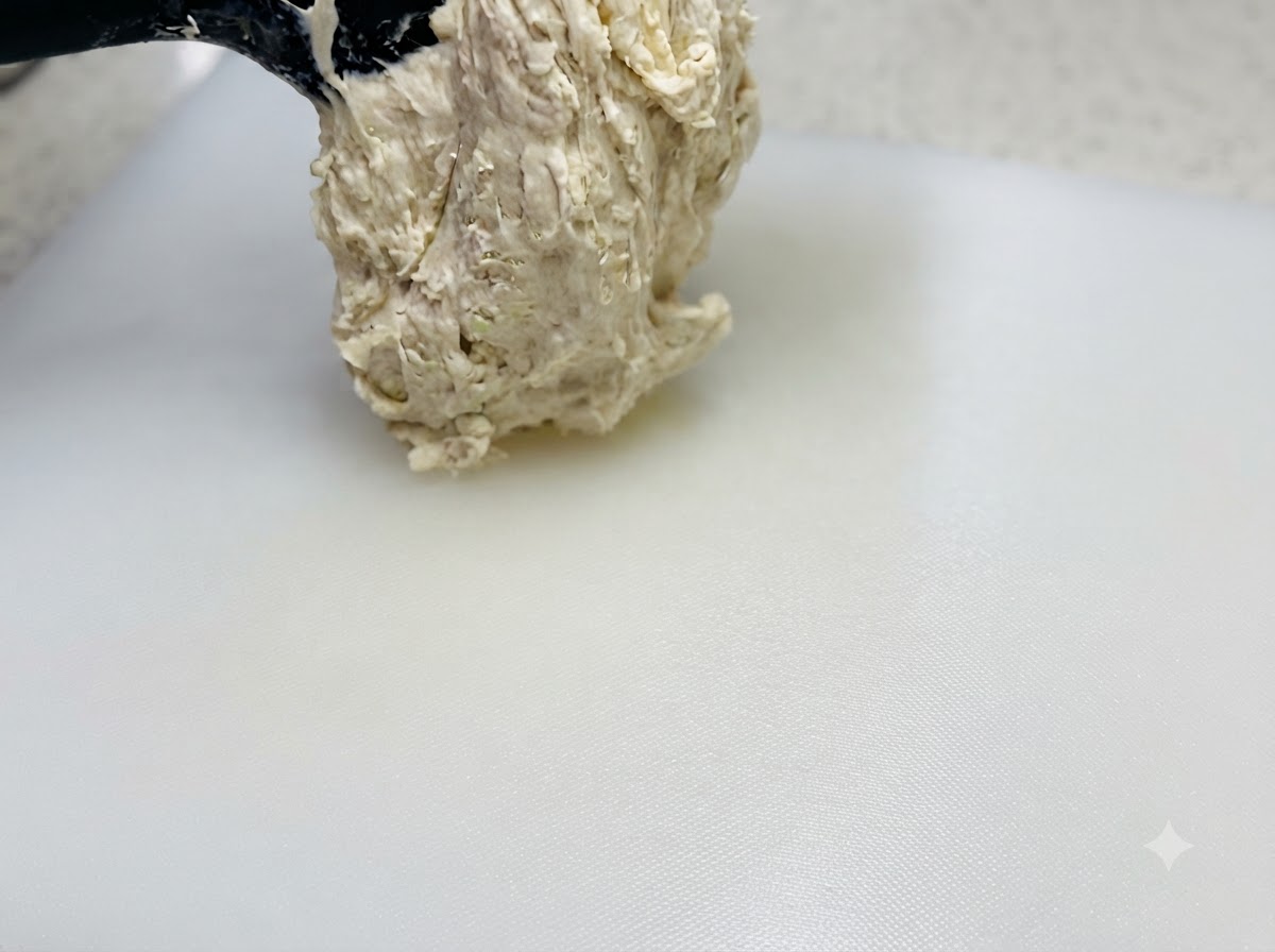 高加水のベタつく生地がスパチュラで持ち上げられ、油がスプレーされた白いカッティングボードの上に置かれている様子。