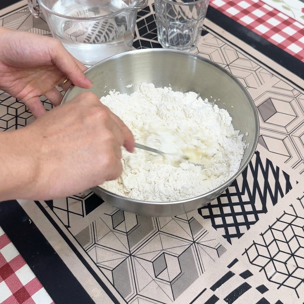 金属製のミキシングボウルの中で、スプーンを使って水と乾いた小麦粉を混ぜ合わせている手。