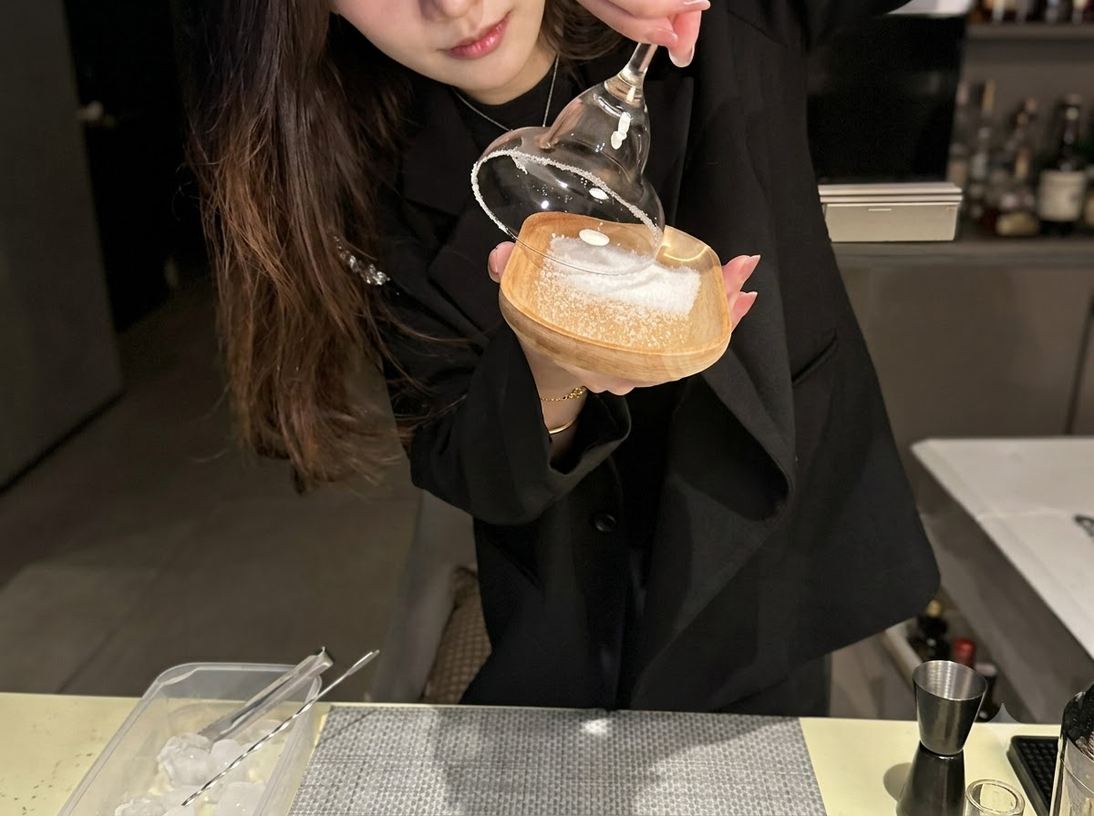 木製の小皿の上に広げられた海塩に、カクテルグラスの縁を押し当てている様子。