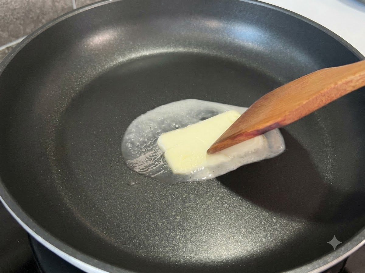 黒いフライパンの中で溶けていく四角いバターを、木製のヘラで軽く押さえている様子。