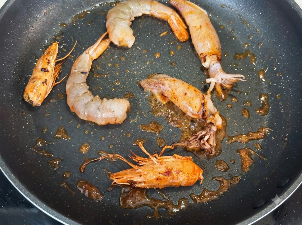 殻をむいたエビ、エビの頭、イカの切り身がフライパンで炒められており、黒こしょうの黒い粒が振られている様子。