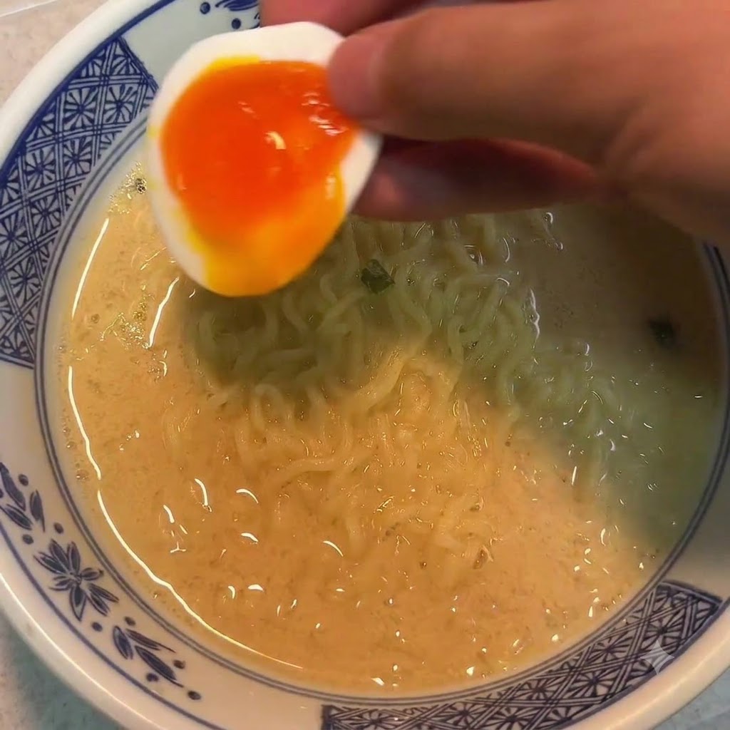 麺の入った器の上に、半分に切った半熟卵を手に持って添えている様子。