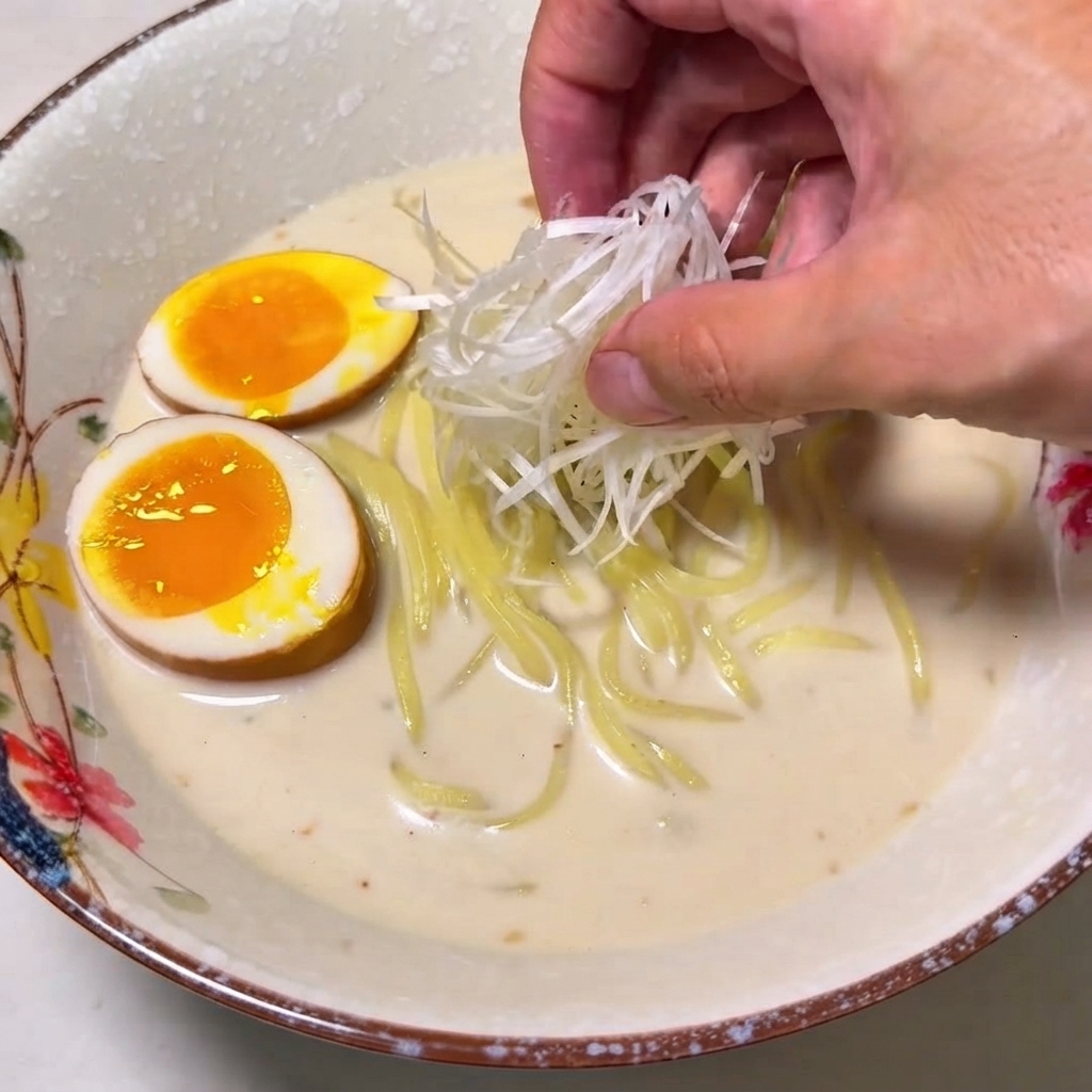ラーメンの丼の中心に、手で白ねぎの千切りの山を盛り付けている様子