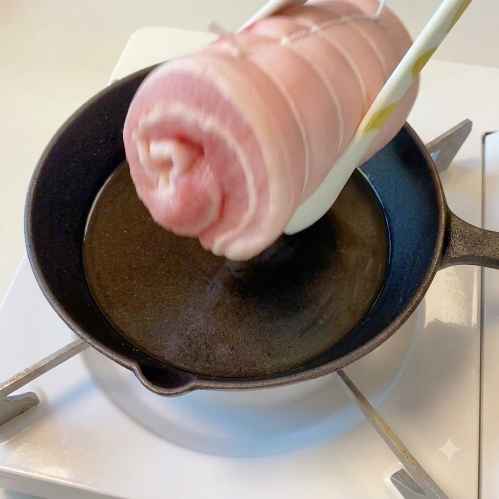 トングで掴まれたタコ糸巻きの豚肉が、油を引いた熱々のフライパンの上にある様子。