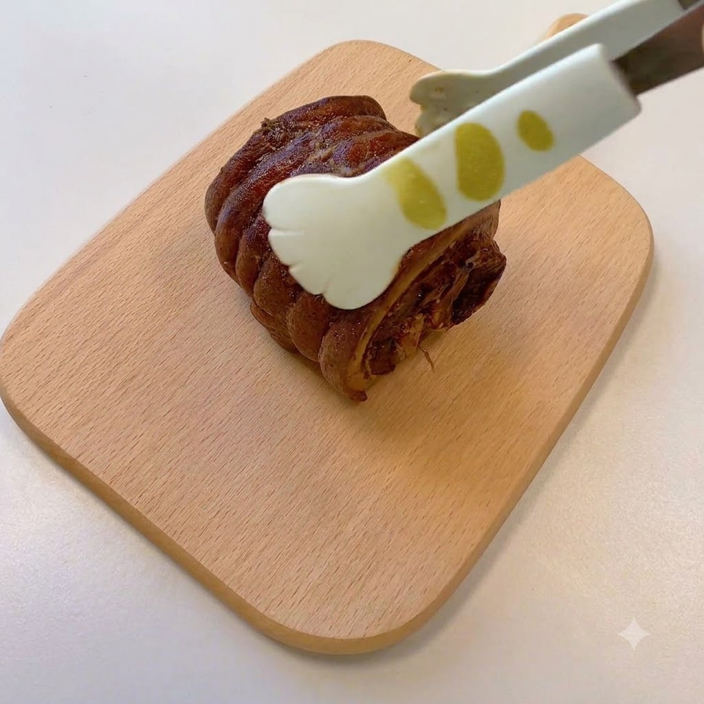 木製のまな板の上で、トングが調理済みのロールチャーシューを掴んでいる様子。