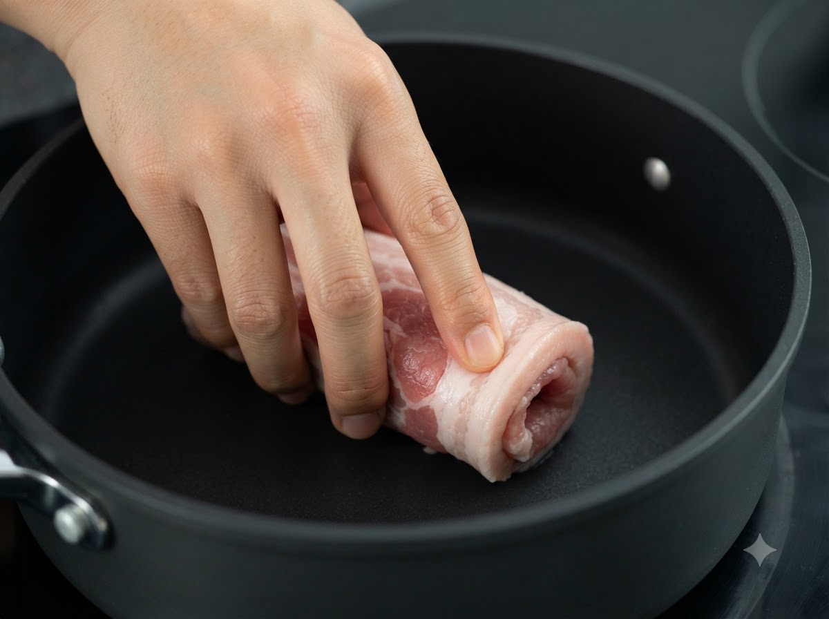 加熱された黒いフライパンの中で焼き目をつけられている、縛られた豚バラ肉のロール。