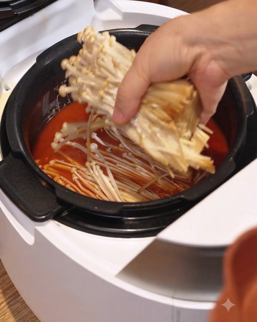 白い電気炊飯器に入った赤いシチューのスープに、えのき茸を入れている様子