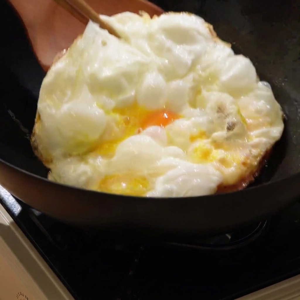 熱い中華鍋の中で、木べらを使って大きなきつね色の目玉焼きを裏返している様子。