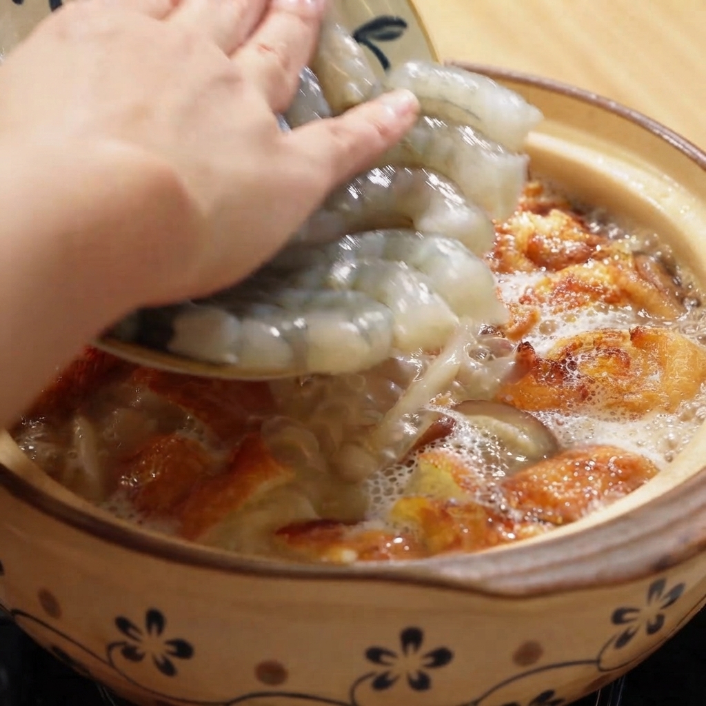 スープと揚げ卵のピースが入った泡立つ土鍋に、生のむき海老を加えている様子。
