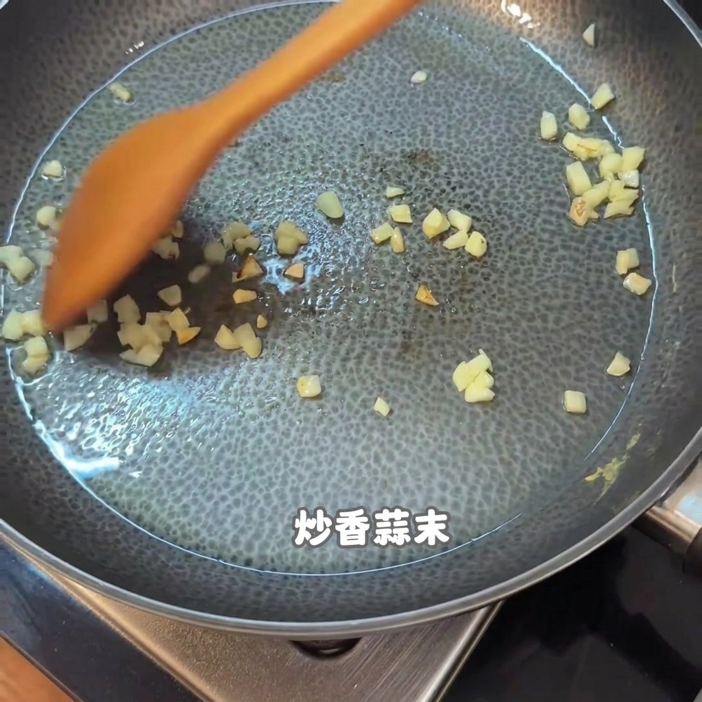 テフロン加工のフライパンの中で、オレンジ色の木製ヘラでかき混ぜられる刻んだにんにくと食材。