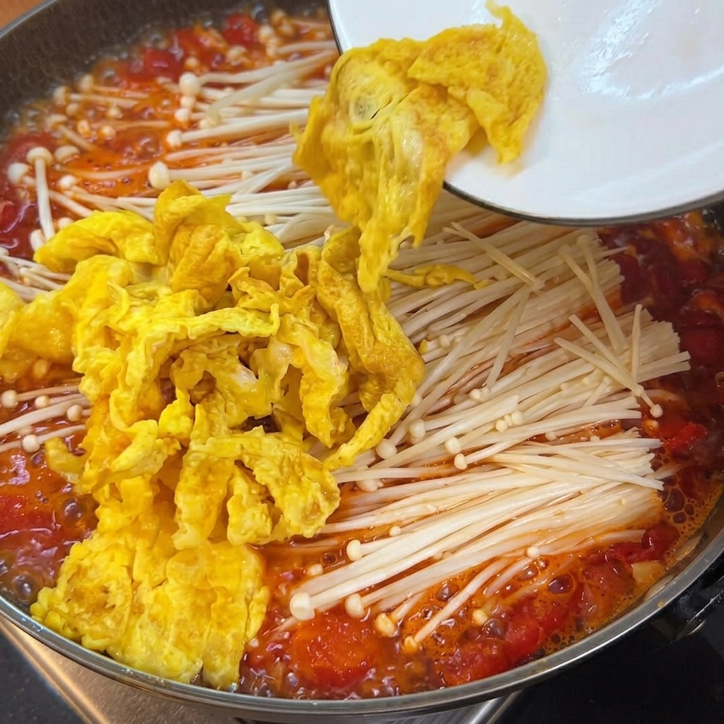 トマトスープ、えのき茸、卵が入った鍋に、なめらかな豆腐の角切りが加えられる様子。