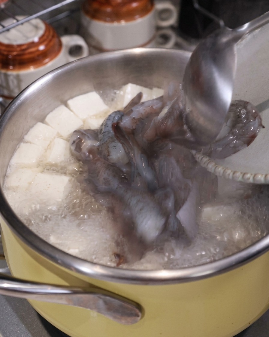 豆腐と豚肉の入った沸騰したスープの鍋に、お玉を使って生のイカとエビの切り身を入れている様子。