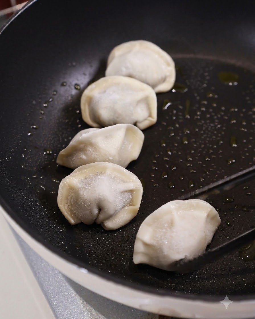 キッチンのトングを使って、油を引いた黒いフラットなフライパンに、他のいくつかの餃子と一緒に生冷凍餃子を置いている様子。