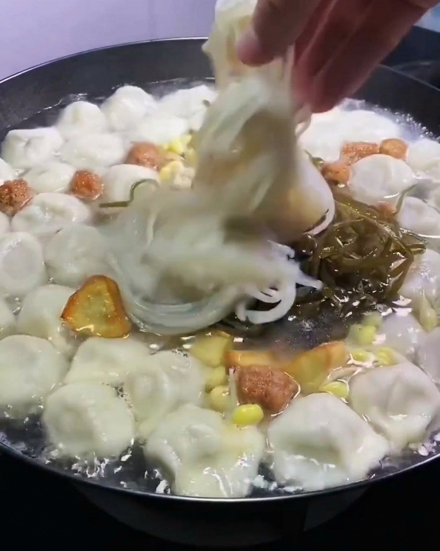 茹で中の餃子とスープの入った鍋に、刻み昆布と半透明の春雨を手で入れているところ。