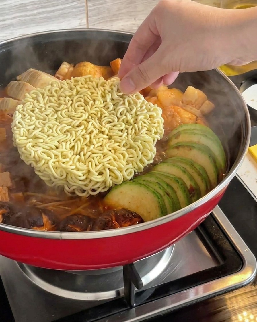 グツグツと煮立つ韓国部隊チゲの中心に、乾麺のインスタントラーメンを入れようとしている手。